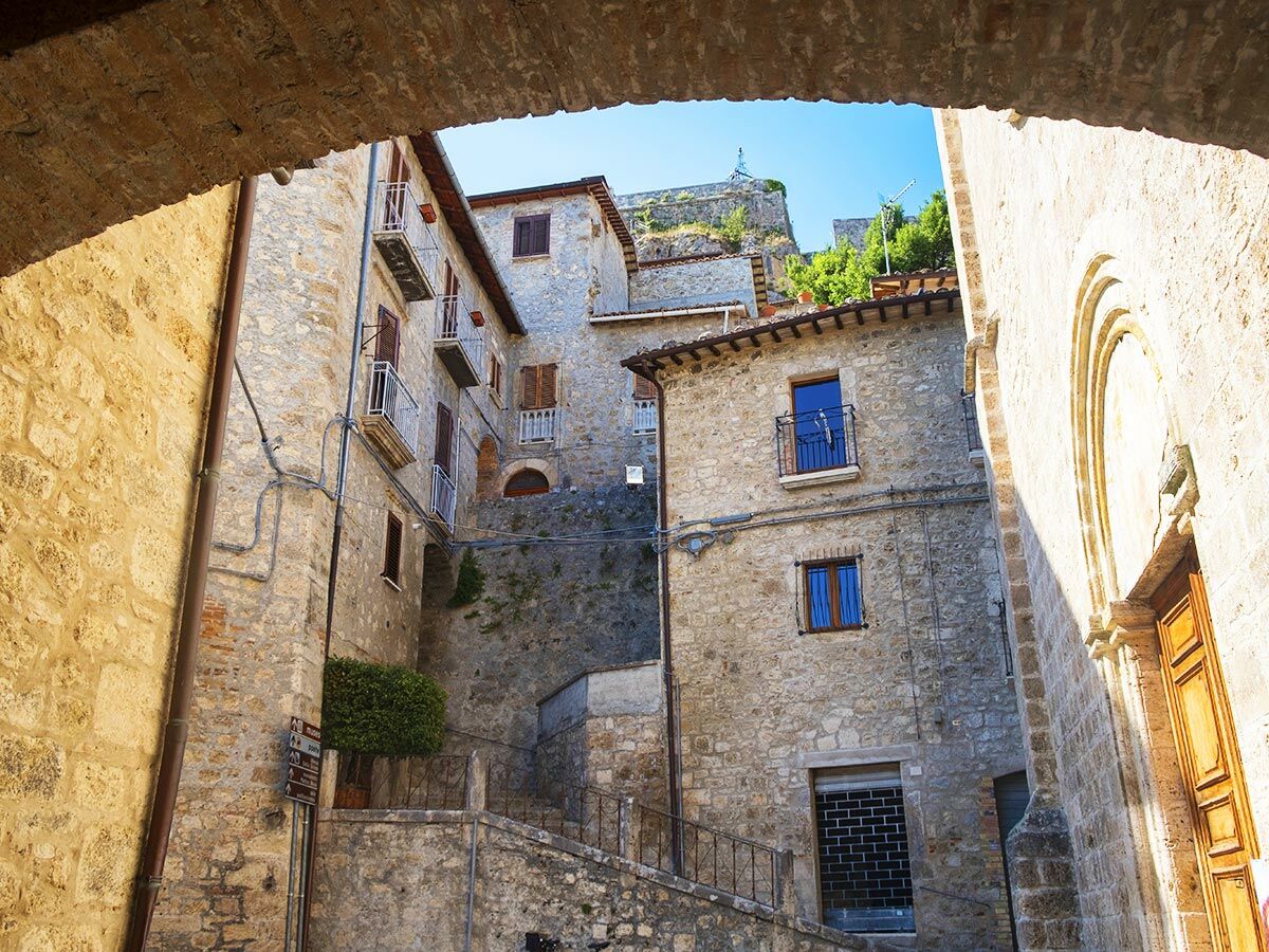 Enge Gasse in historischer italienischer Altstadt, umrahmt von Steinbauten, Balkonen und Treppen, blauer Himmel und Burgblick.