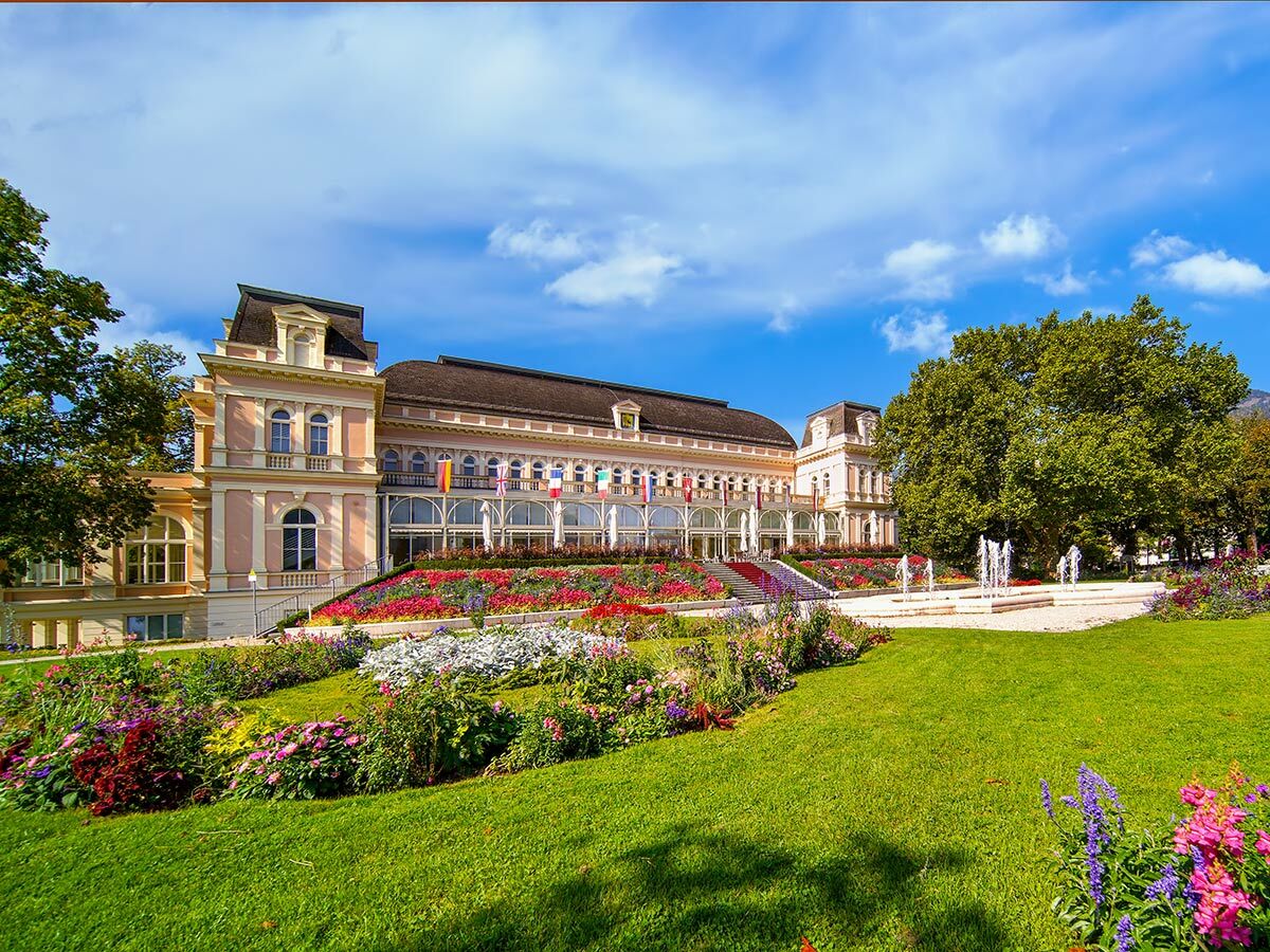Elegantes Kurhaus mit farbenfrohen Blumenbeeten, grünen Rasenflächen und Springbrunnen unter blauem Himmel.