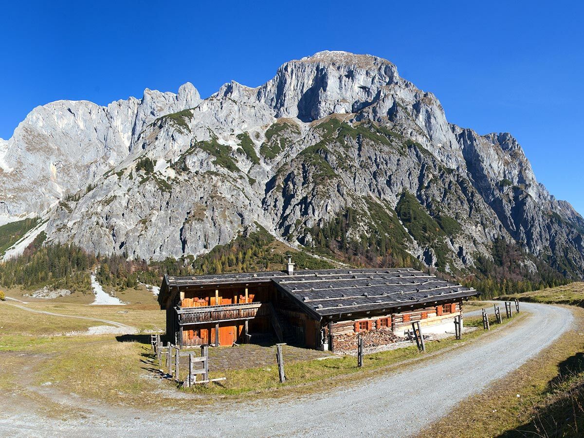 Rustikale Holzhütte mit Holzstapel und Balkon vor majestätischen Alpenbergen unter strahlend blauem Himmel, ein Schotterweg führt vorbei.