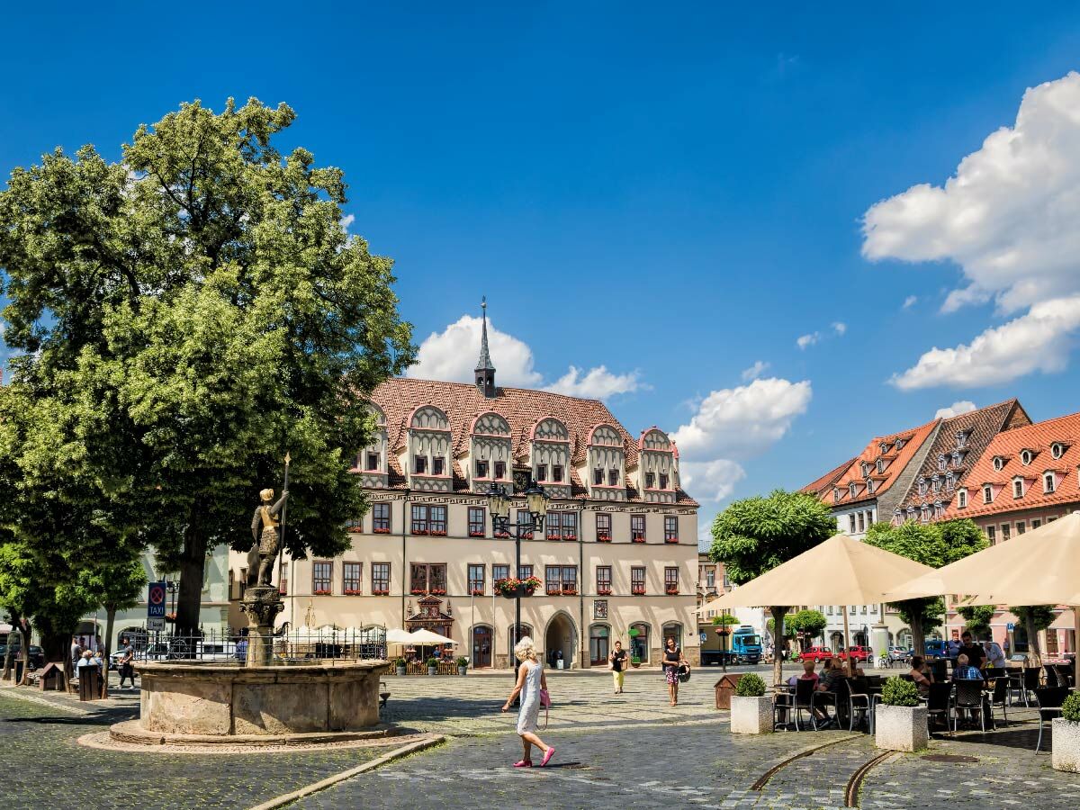 Belebtes historisches Zentrum: Rathaus, Rolandbrunnen, Baum, Cafés auf Kopfsteinpflasterplatz unter blauem Himmel.