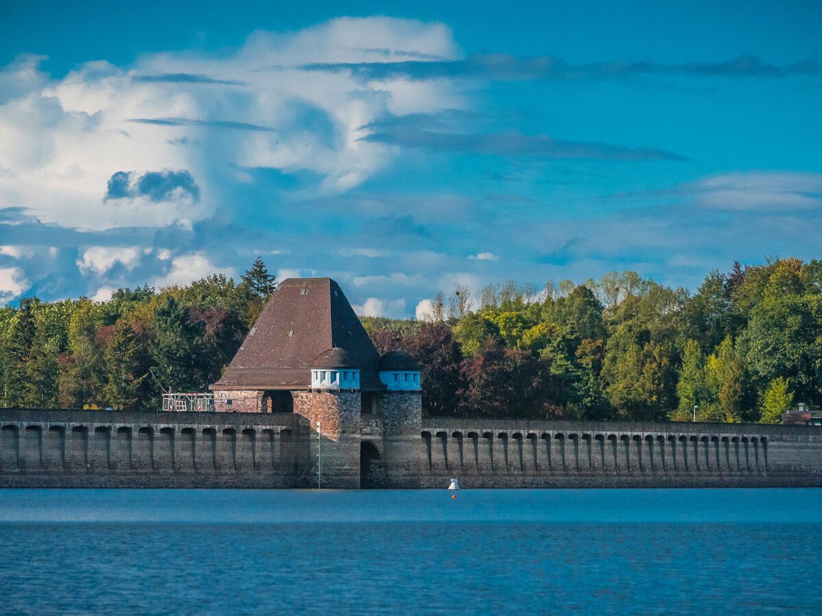 Historisches Dammgebäude mit markantem Dach und Türmen am Edersee, umgeben von Wald unter blauem Himmel mit Wolken.