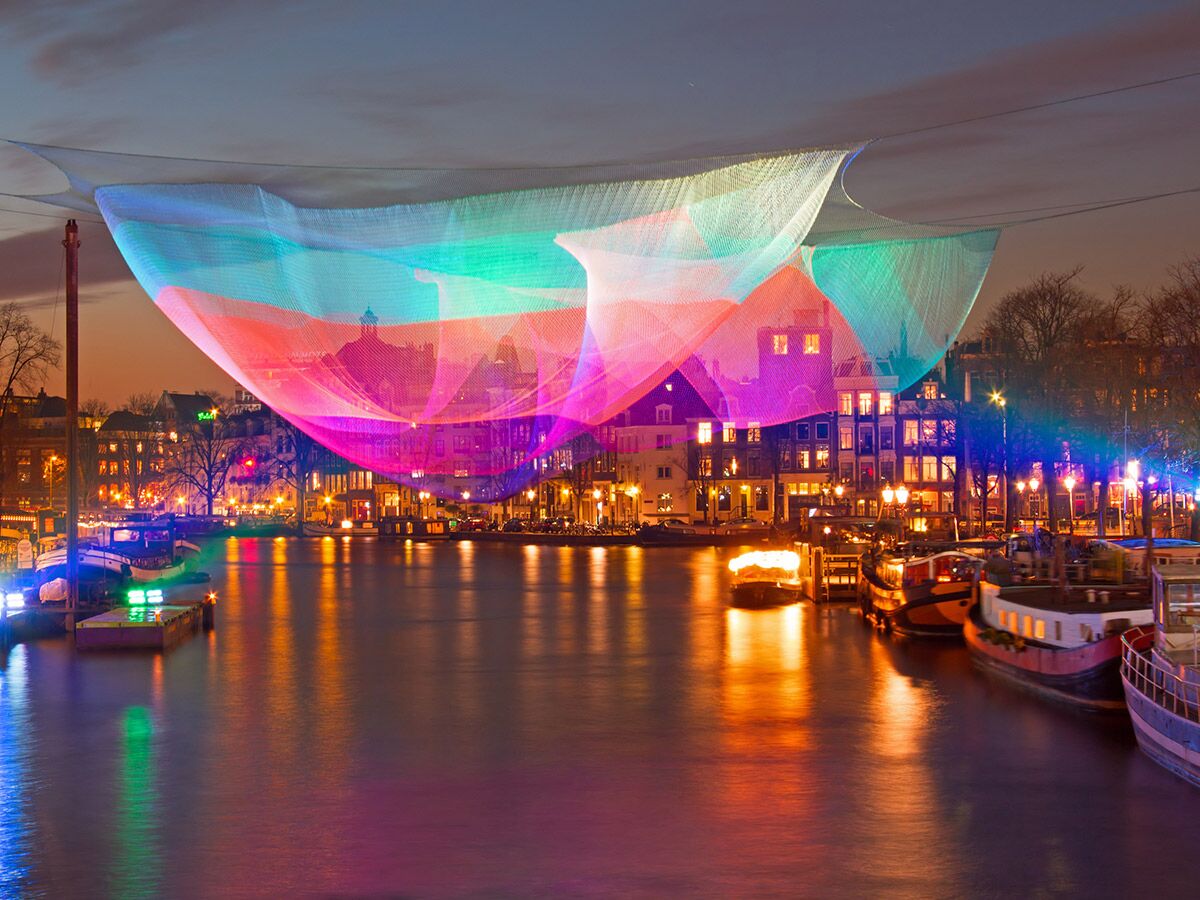Eine farbenfrohe Lichtkunstinstallation aus Netzen spannt sich nachts über einen beleuchteten Stadtkanal mit Booten und historischen Gebäuden.