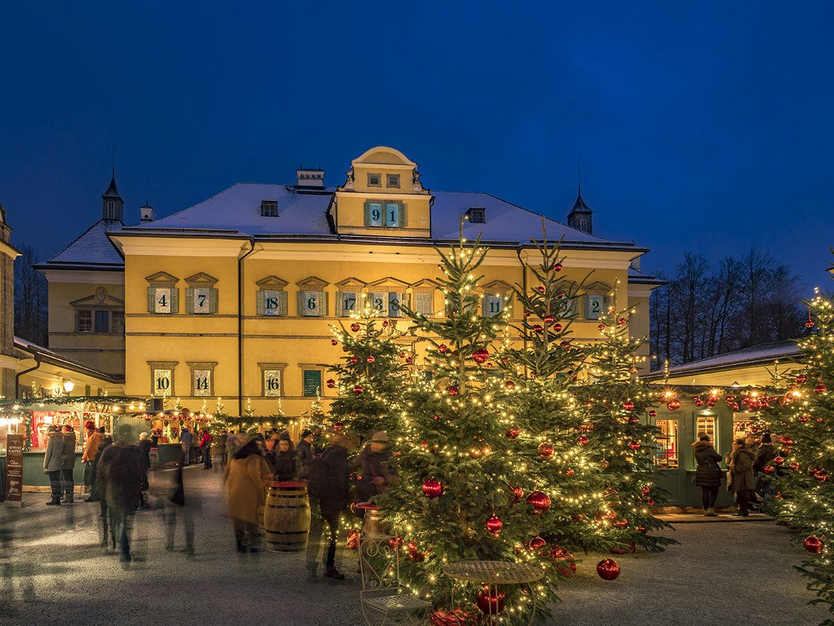 Weihnachtsmarkt Hellbrunn Schloss bei Dämmerung, mit Adventkalender-Fenstern, festlich beleuchteten Bäumen und Besuchern.