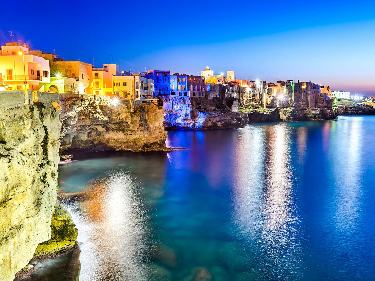 Polignano a Mare in Apulien bei Nacht: Beleuchtete Häuser auf Klippen am tiefblauen Meer mit hellen Lichtreflexionen.