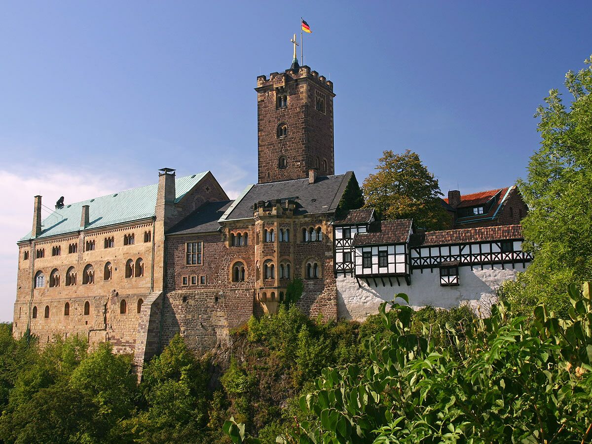 Die historische Wartburg in Deutschland, ein majestätisches Schloss mit Turm, umgeben von grünen Bäumen und Fachwerkhäusern.
