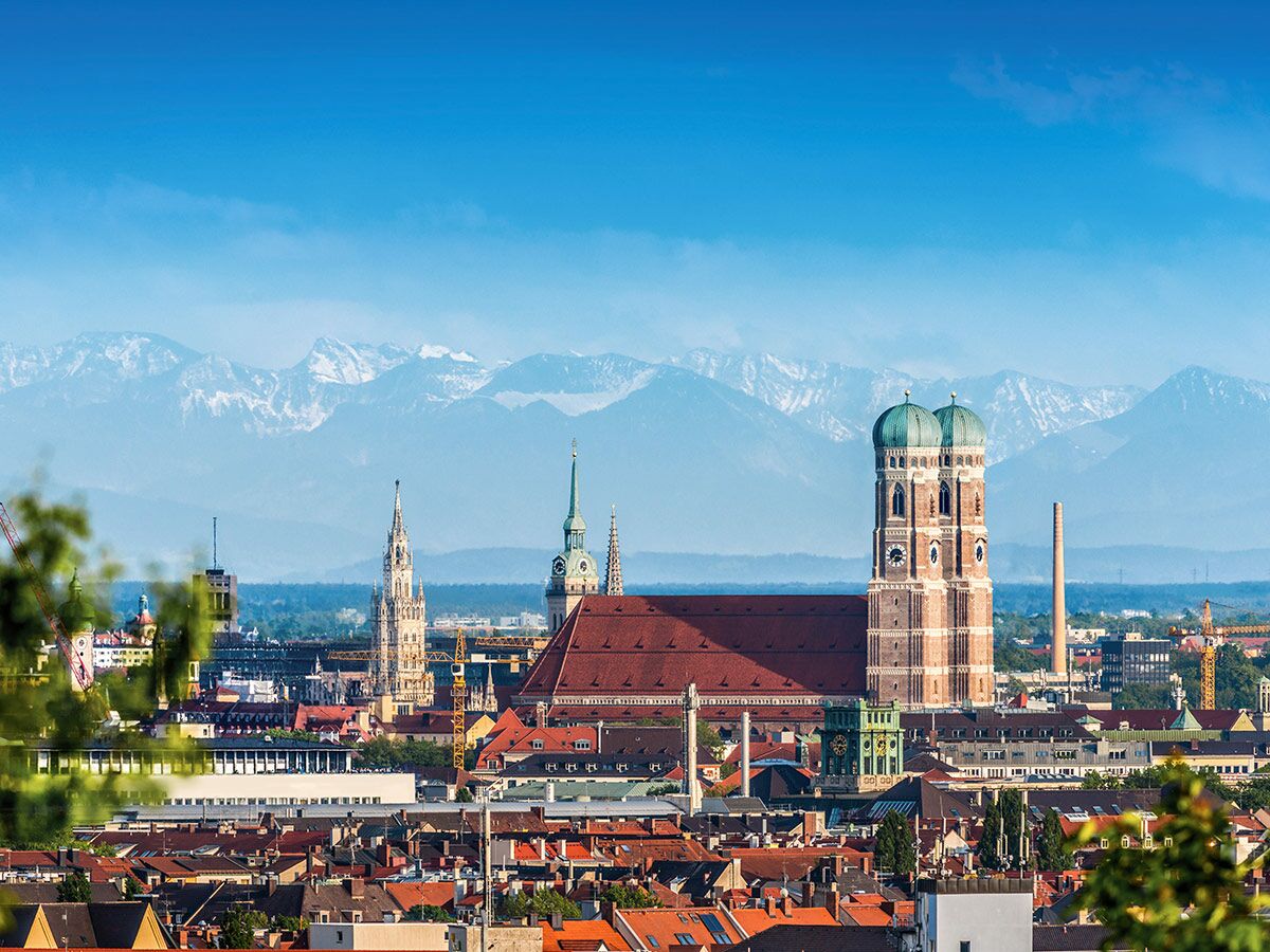 Panoramablick auf München mit Frauenkirche, roten Dächern und schneebedeckten Alpen im Hintergrund bei blauem Himmel.