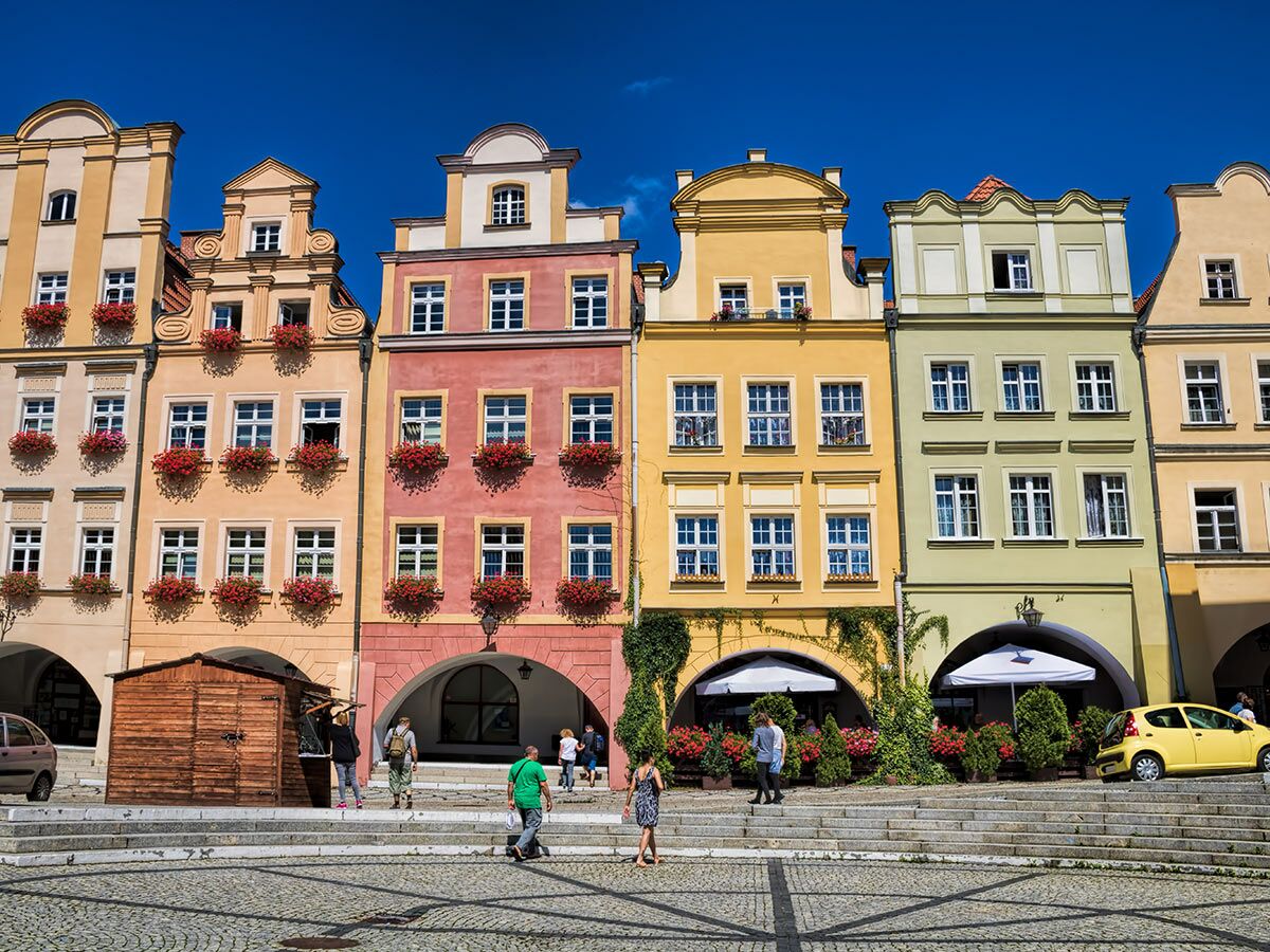 Reihe bunter historischer Häuser mit Blumenkästen und Arkaden an einem sonnigen Marktplatz. Menschen gehen über das Kopfsteinpflaster.