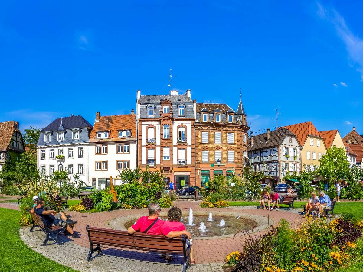 Idyllischer Marktplatz mit historischen Gebäuden, sprudelndem Brunnen und entspannten Menschen auf Bänken bei Sonnenschein.