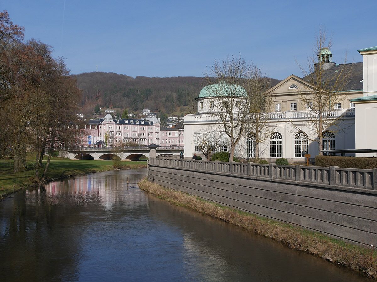 Flusslandschaft in Bad Ems mit der Lahn, historischen Gebäuden wie dem Kurhaus und dem Häcker's Grand Hotel, einer Brücke und bewaldeten Hügeln unter blauem Himmel.
