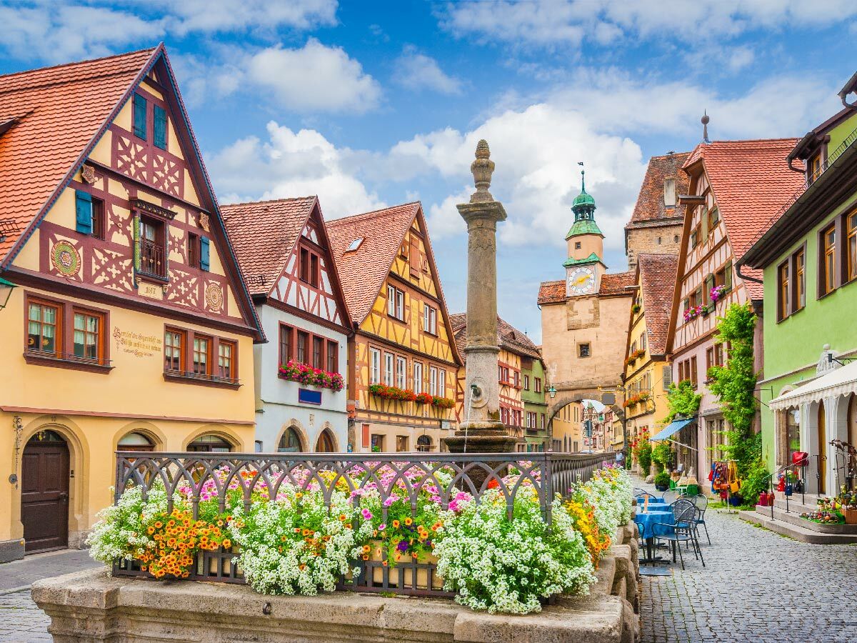 Malerische Gasse in Rothenburg ob der Tauber mit bunten Fachwerkhäusern, Brunnen mit Blumen und historischem Uhrturm unter blauem Himmel.