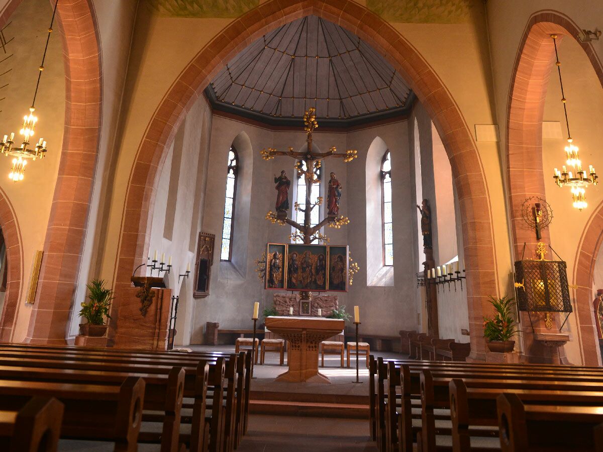 Ansicht eines historischen Kircheninnenraums mit zentralem Altar, Holzbänken, hohen Gewölben und beleuchteten Kronleuchtern.