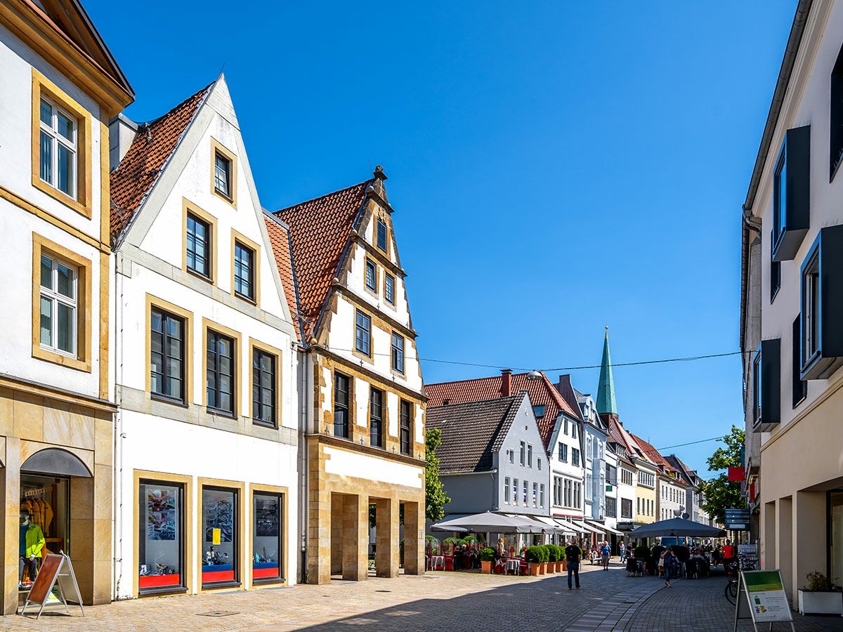 Blick auf eine belebte Fußgängerzone in einer europäischen Altstadt mit historischen Gebäuden, Geschäften und Straßencafés unter blauem Himmel.