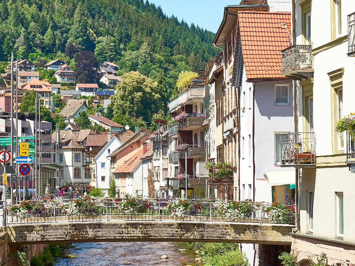 Malerisches Dorf im Schwarzwald mit blumengeschmückter Brücke über einen Fluss und Häusern am bewaldeten Hang bei Sonnenschein.