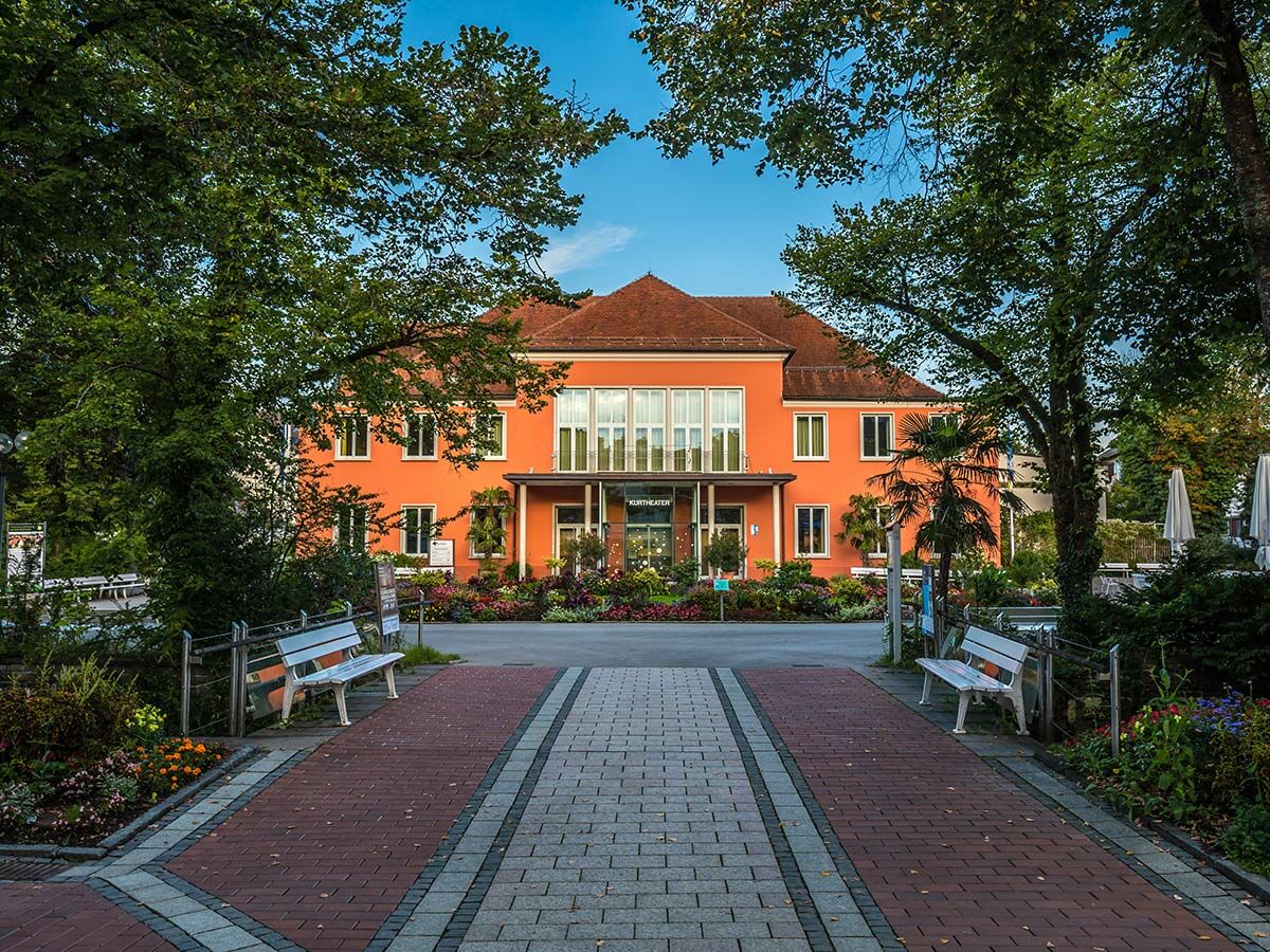 Orangefarbenes Kurtheatergebäude umgeben von üppigem Grün und Bäumen, mit gepflastertem Weg und Bänken davor unter blauem Himmel.