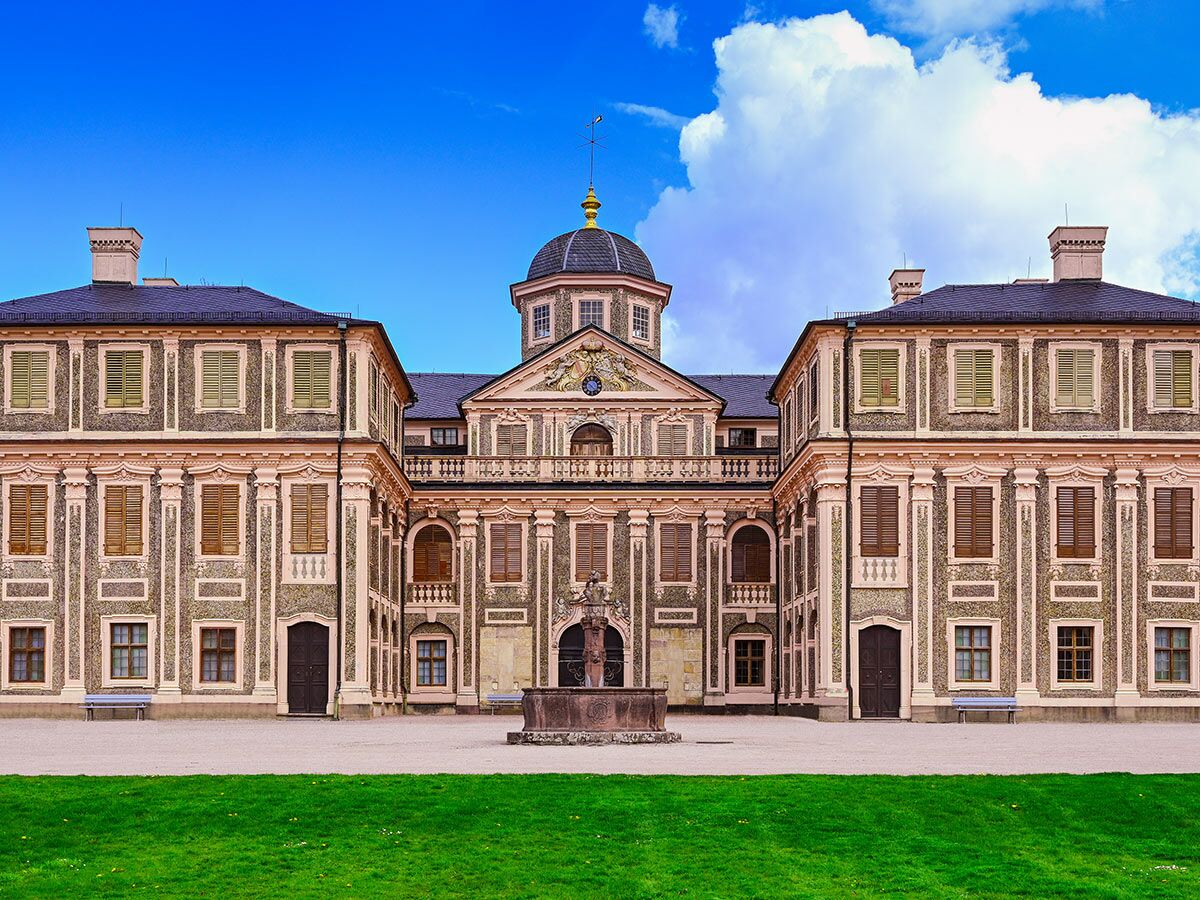 Majestätisches Barockschloss mit Kuppel, aufwendiger Fassade, Innenhof mit Steinbrunnen und grünem Rasen unter blauem Himmel.