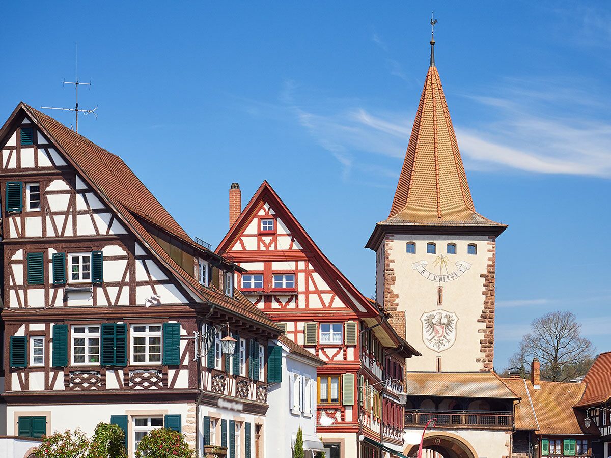 Historische Fachwerkhäuser mit grünen Fensterläden und ein Stadttor-Turm mit Sonnenuhr und Wappen in einer deutschen Altstadt bei blauem Himmel.