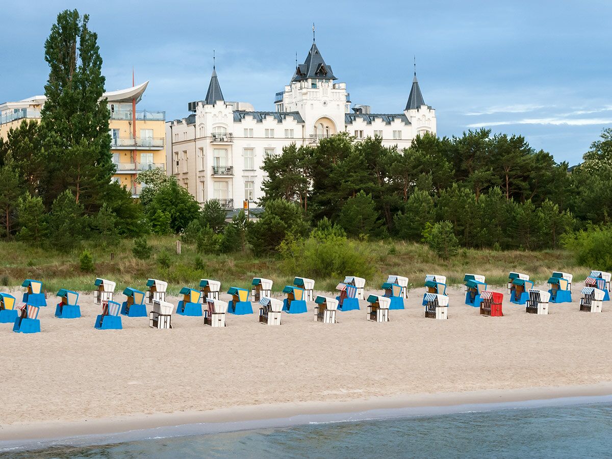 Reihen von Strandkörben auf einem Sandstrand am Meer mit bewaldeter Dünenlandschaft und imposanten Hotels im Hintergrund.