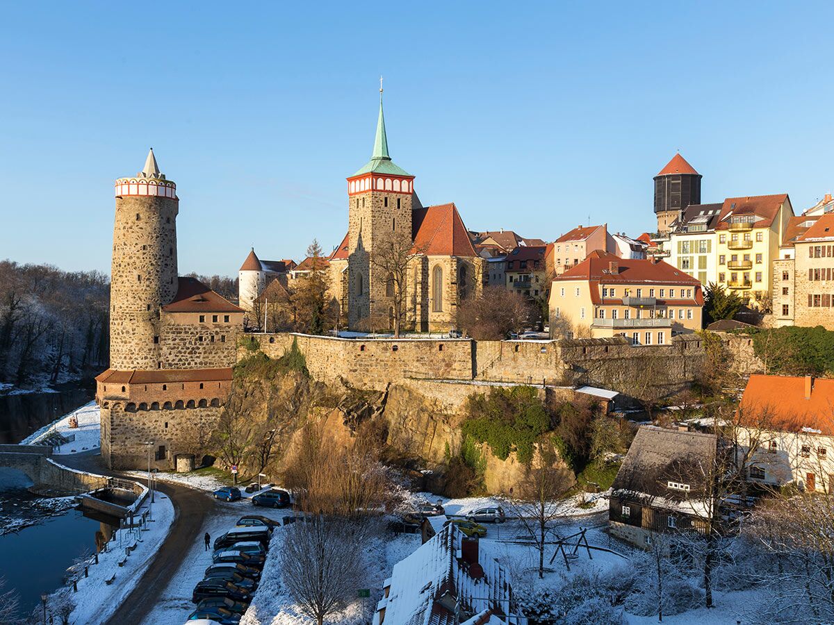 Historische Ortenburg Bautzen im Winter, mit Schnee und Spreeufer im Vordergrund unter blauem Himmel.