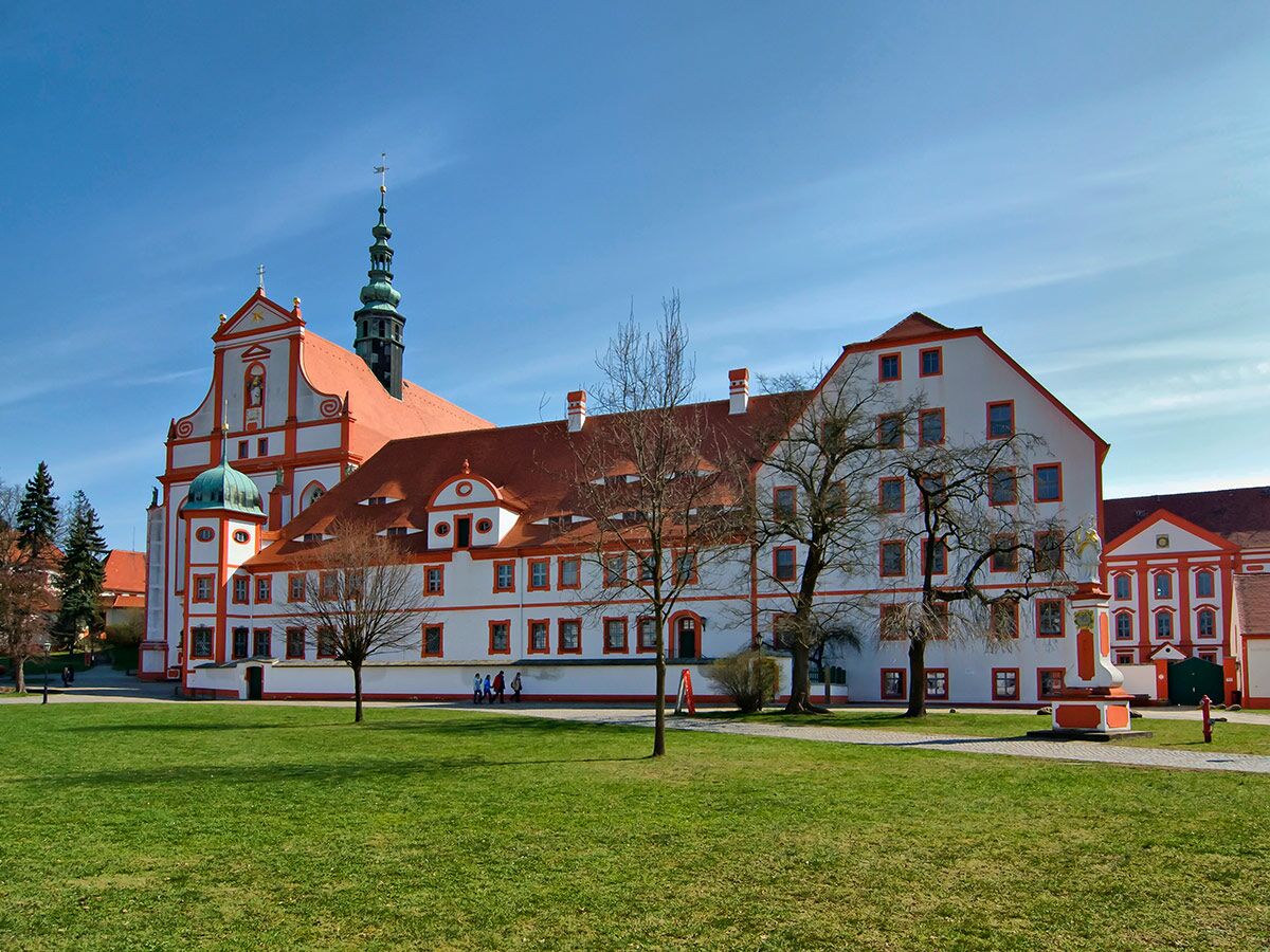 Barockes Klostergebäude mit Kirche, Turm und roten Dächern unter blauem Himmel, grüne Wiese im Vordergrund.