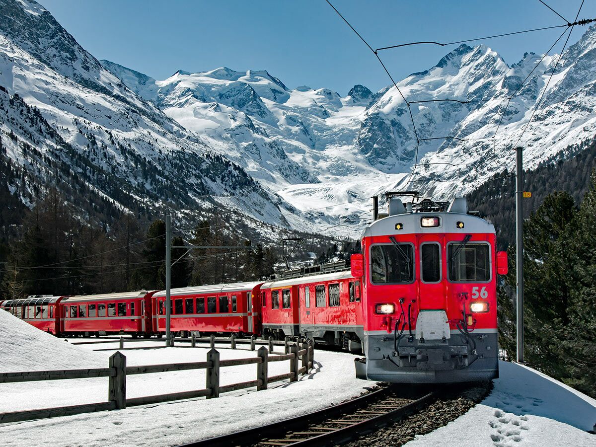 Eine rote Bergbahn fährt auf Gleisen durch eine verschneite Alpenlandschaft mit hohen Bergen und Bäumen unter blauem Himmel.