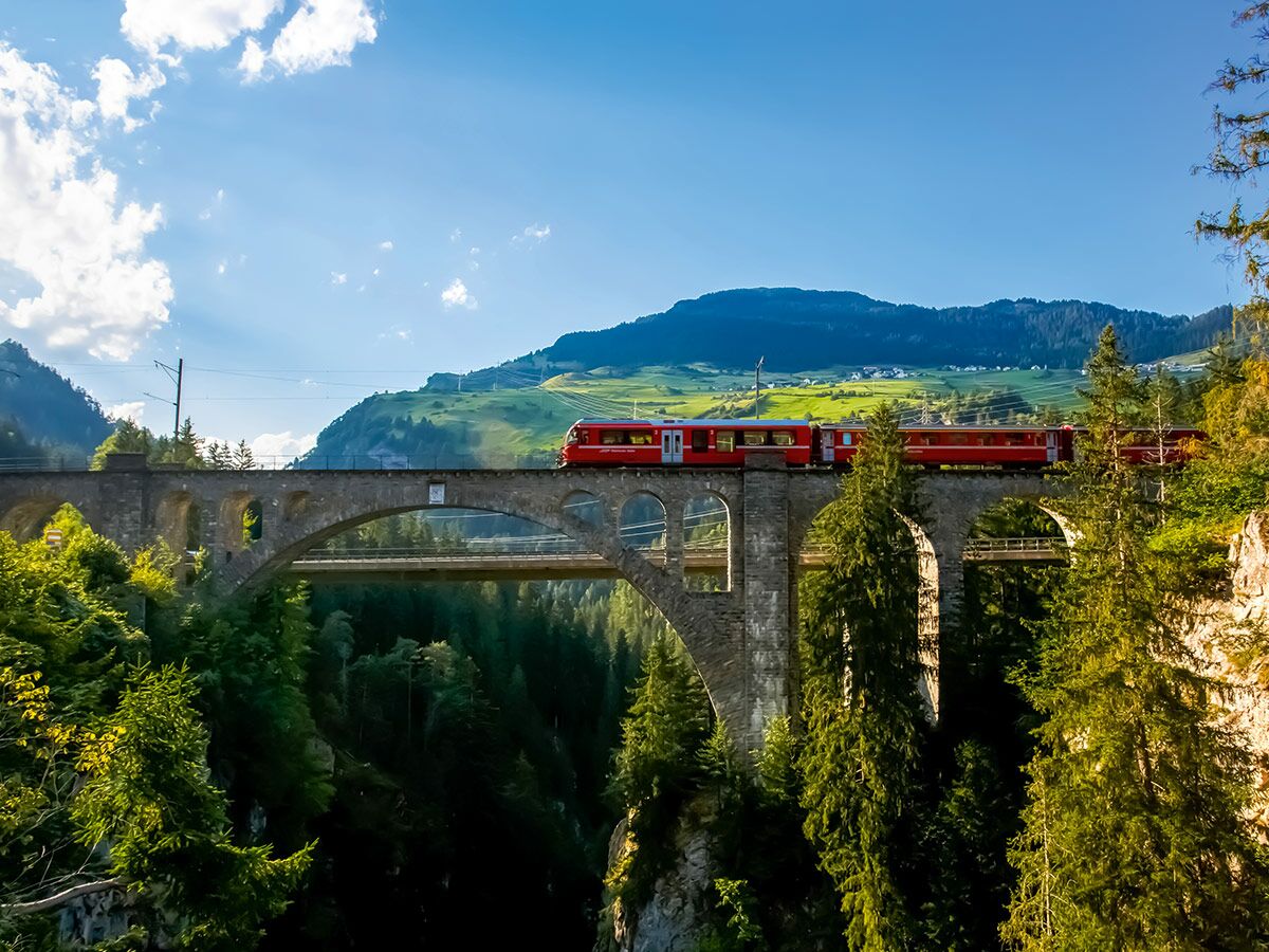 Roter Zug der Rhätischen Bahn überquert ein historisches Steinviadukt in den sonnigen Schweizer Alpen.