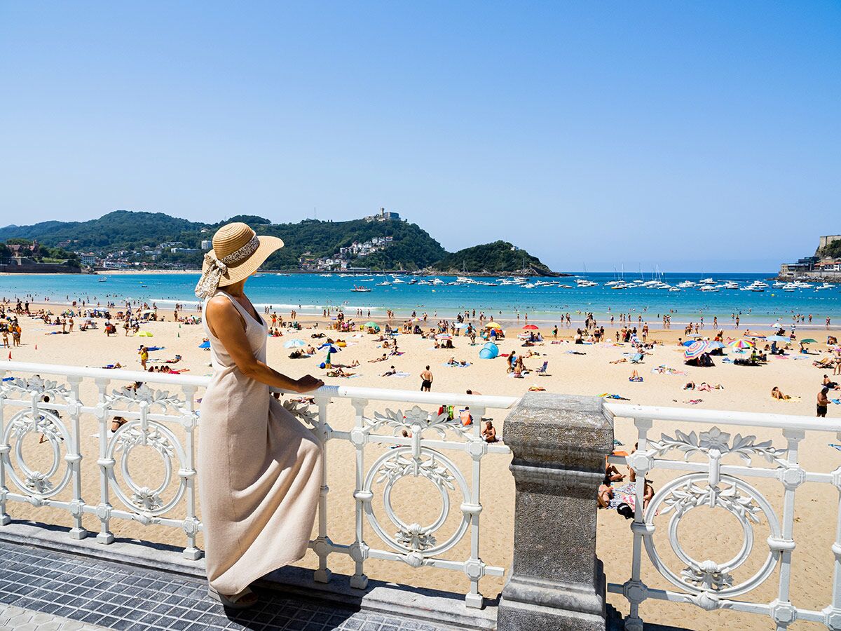 Frau mit Strohhut und Kleid blickt auf den belebten Strand von San Sebastián mit Badegästen und Booten an einem Sommertag.