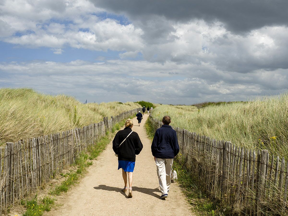 Zwei Personen gehen auf einem Sandweg, umgeben von grasbewachsenen Dünen und Holzzaun, unter bewölktem Himmel.