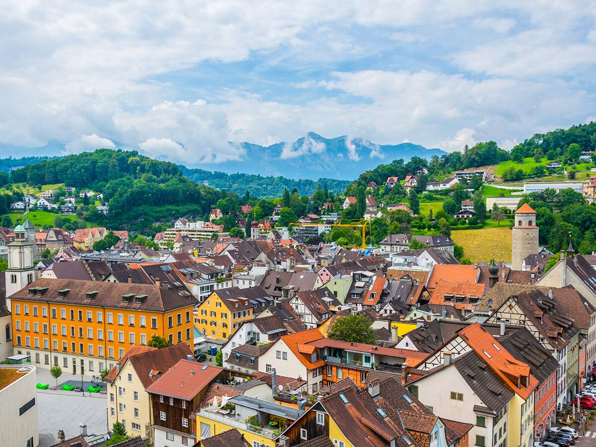 Luftaufnahme einer malerischen historischen Stadt in den Alpen mit bunten Häusern, Bergen und grünen Hügeln unter bewölktem Himmel.