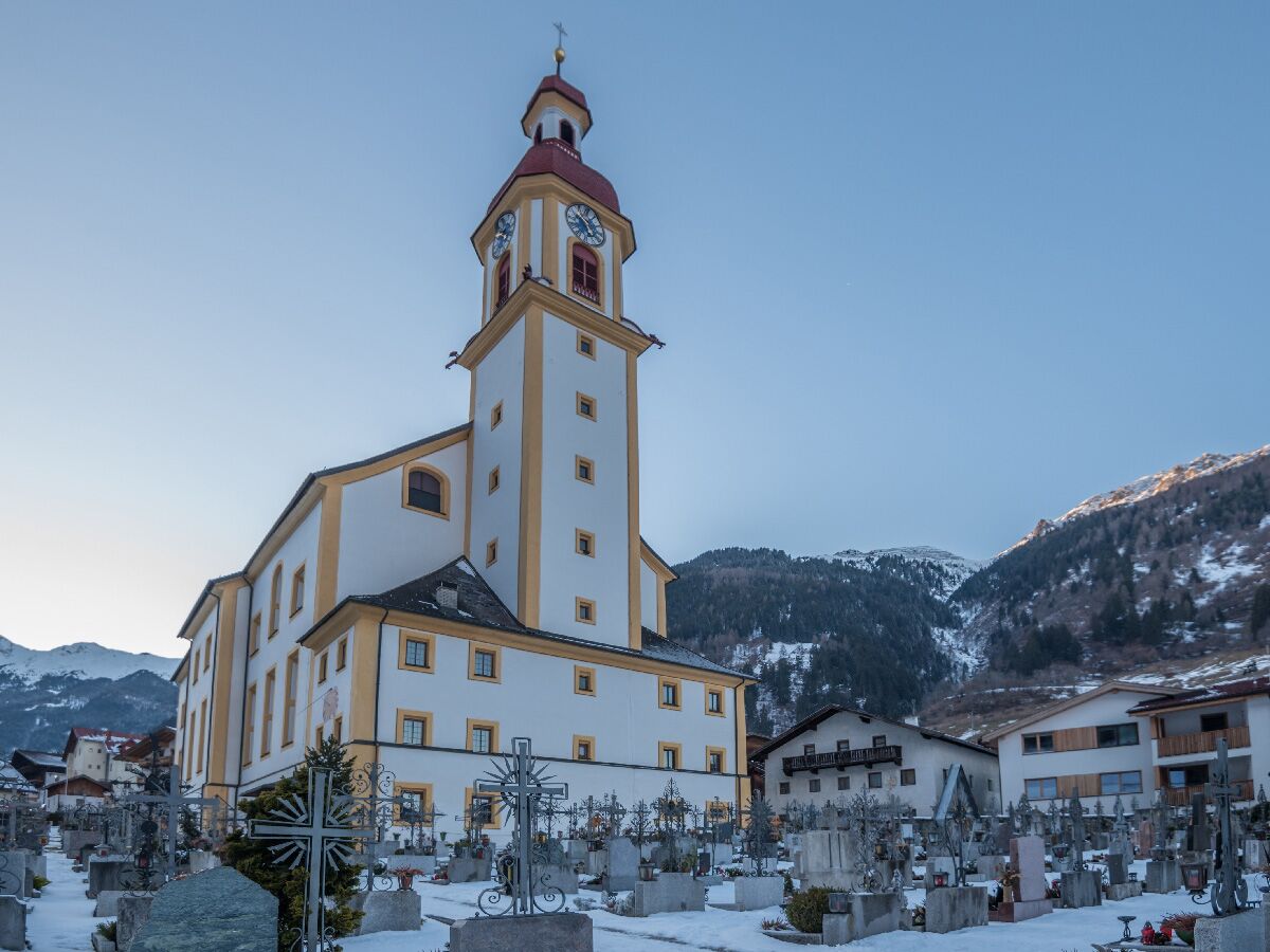 Barocke Kirche mit Glockenturm und verschneitem Friedhof im Alpenpanorama, umgeben von Häusern und Bergen.