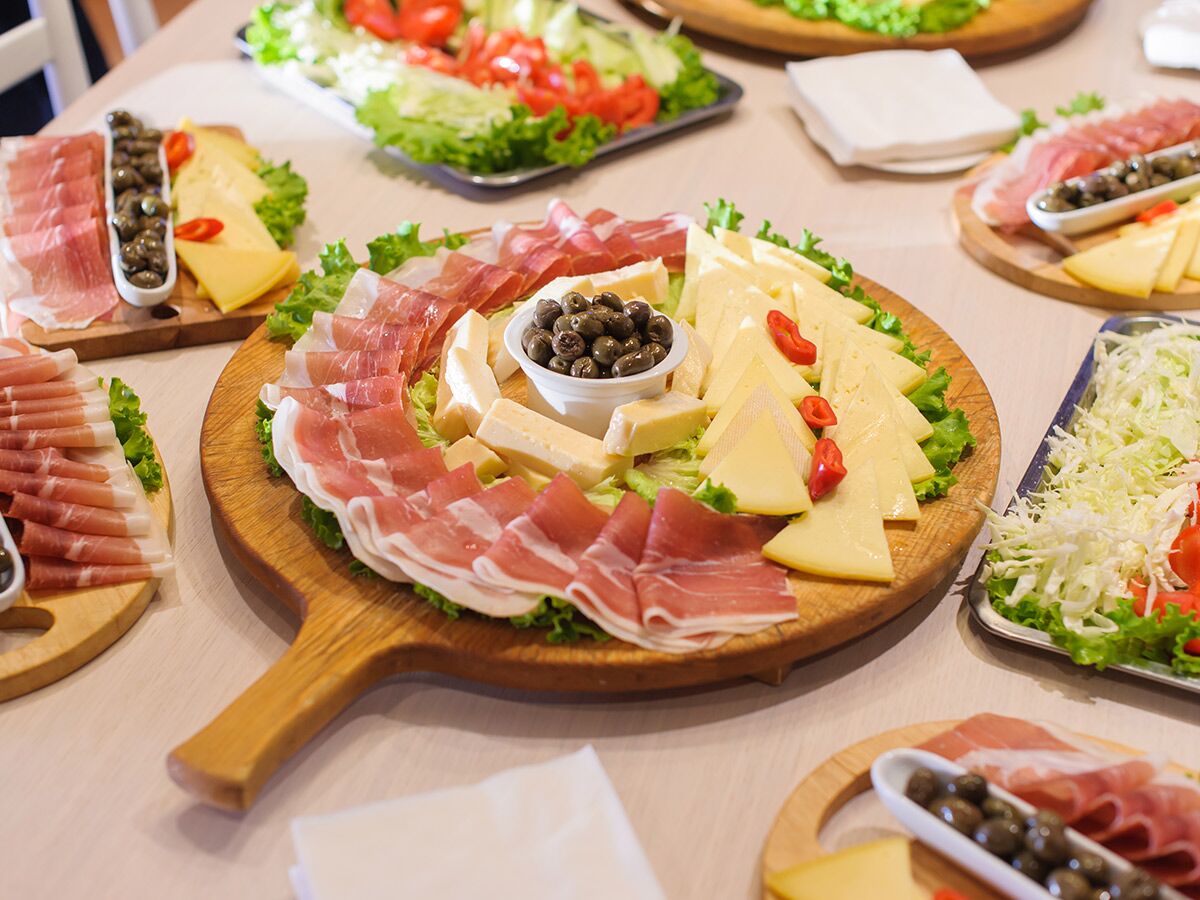 Reichhaltiges Delikatessen-Buffet mit großen Platten voller Schinken, Käse, Oliven, und frischem Salat auf einem Holztisch.
