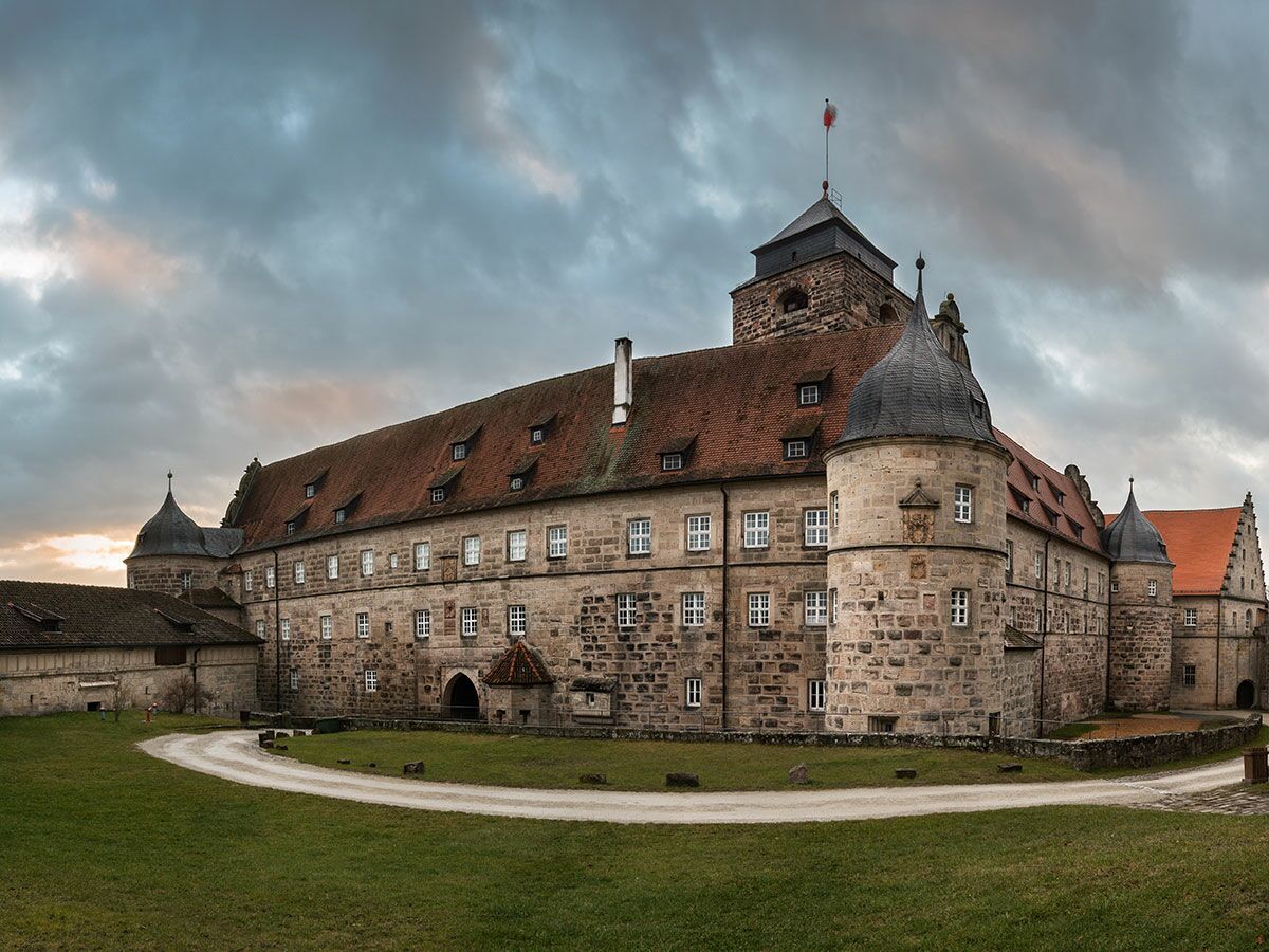Große historische Burg aus Stein mit roten Dächern und mehreren Türmen unter dramatischem Wolkenhimmel bei Sonnenuntergang.