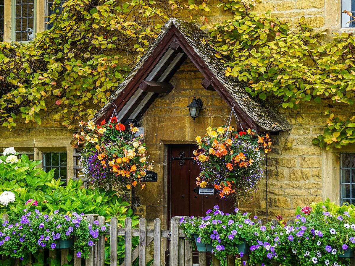 Eingang eines charmanten Steincottages mit bunten Blumen in Hängekörben, grünem Laubwerk und Holzzaun im Vordergrund.