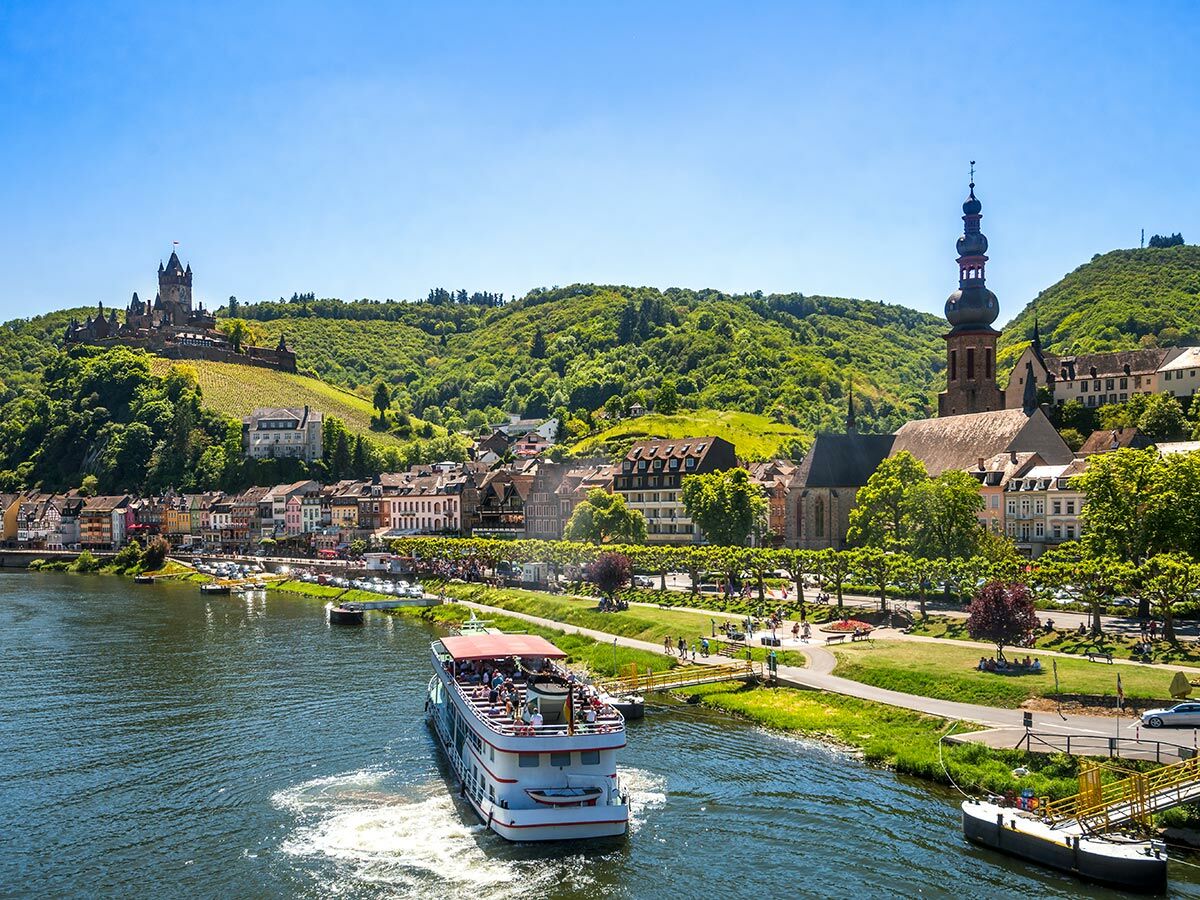 Panoramablick auf Cochem an der Mosel mit Reichsburg, Stadt und Passagierschiff unter blauem Himmel.