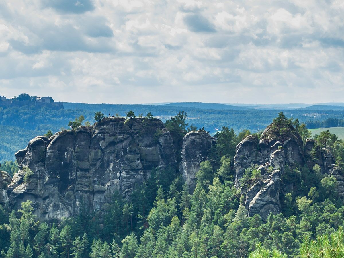 Malerische Felsformationen im Wald der Sächsischen Schweiz mit Burg Königstein am Horizont unter wolkigem Himmel.