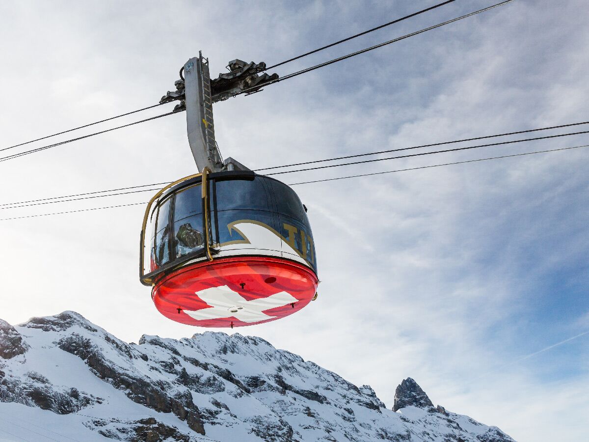 Eine rote Schweizer Gondelbahn mit Flagge schwebt über verschneiten Berggipfeln unter einem bewölkten Himmel.