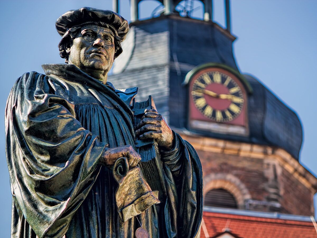 Bronzestatue von Martin Luther, der Schriftrolle und Buch hält, vor einem Kirchturm mit Uhr unter blauem Himmel.