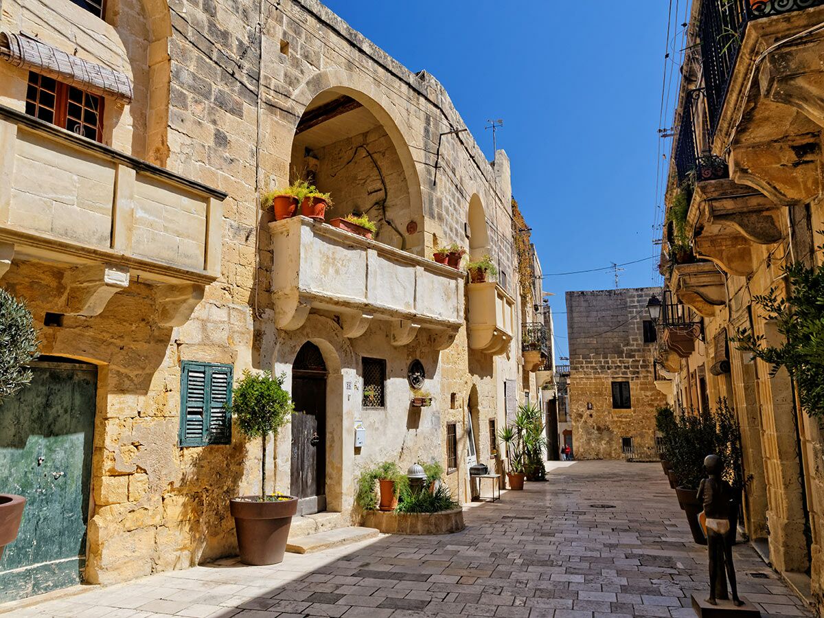 Enge Gasse in Malta mit historischen Steingebäuden, Bögen, Balkonen, Topfpflanzen und klarem blauem Himmel an einem sonnigen Tag.