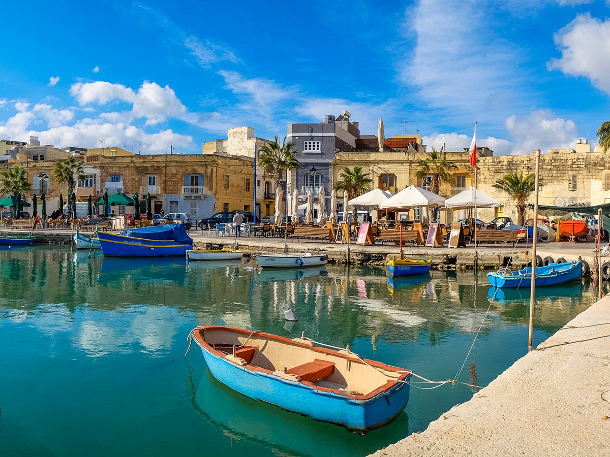 Bunte Fischerboote (Luzzus) im sonnigen Hafen von Marsaxlokk, Malta, mit historischen Gebäuden und Cafés am Ufer.