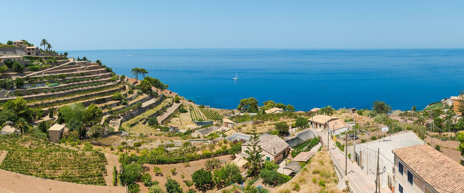 Panoramablick auf eine sonnige Mittelmeerküste mit terrassierten Hängen, grünen Weinbergen, traditionellen Häusern und blauem Meer mit Segelboot.