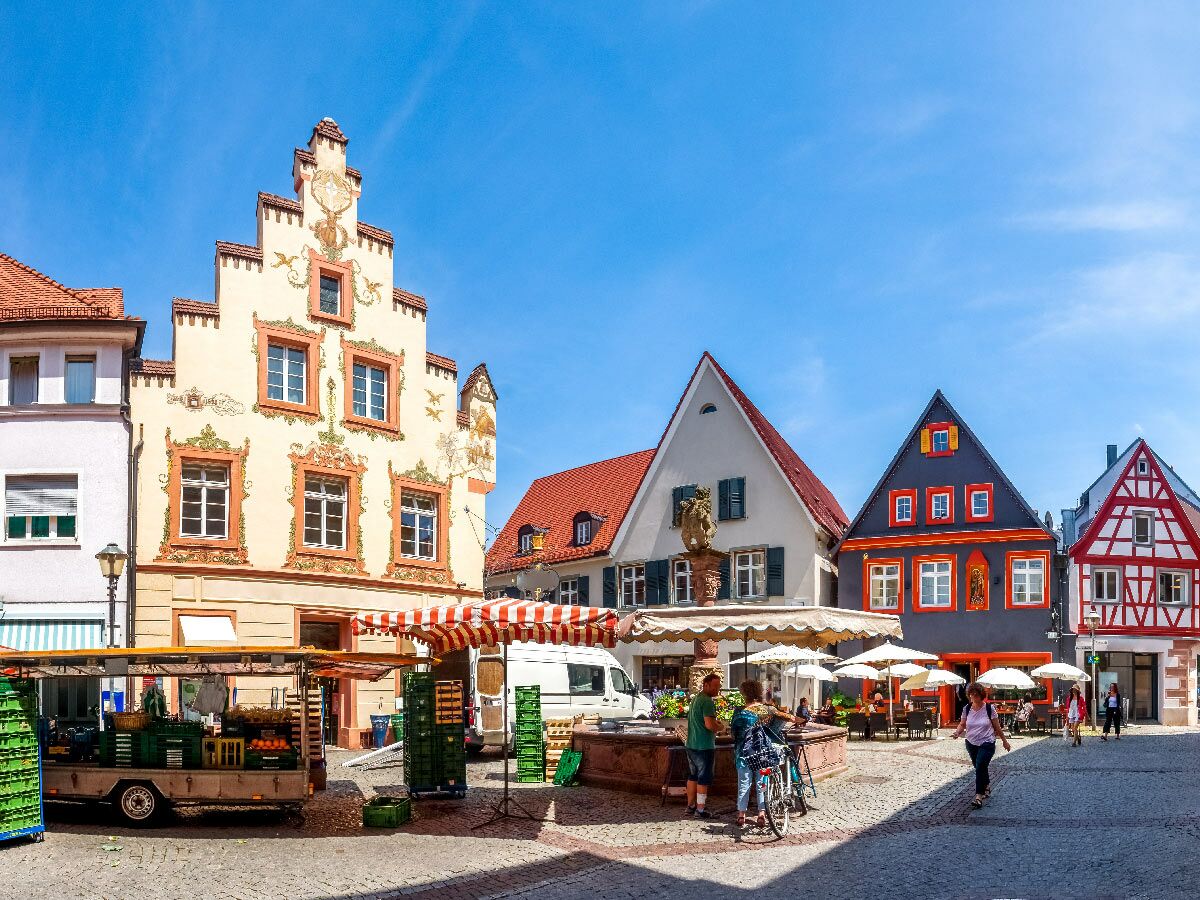 Belebtes Stadtplatz-Panorama in Deutschland: Bunte Giebelhäuser, Marktstände, Brunnen, Cafés und flanierende Menschen unter blauem Himmel.