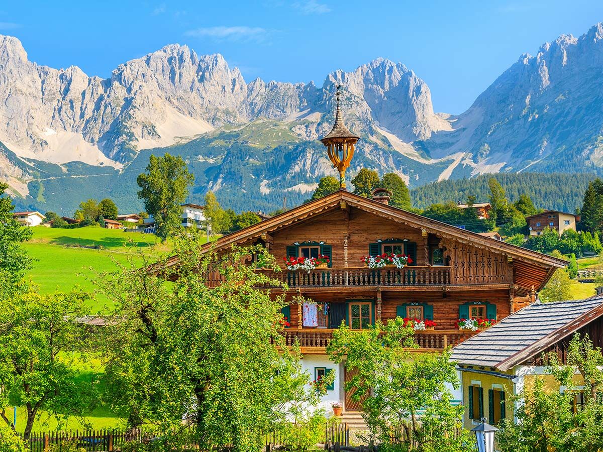 Traditionelles Alpenchalet aus Holz mit Blumenbalkonen, grünen Wiesen und majestätischen Bergen unter blauem Himmel.
