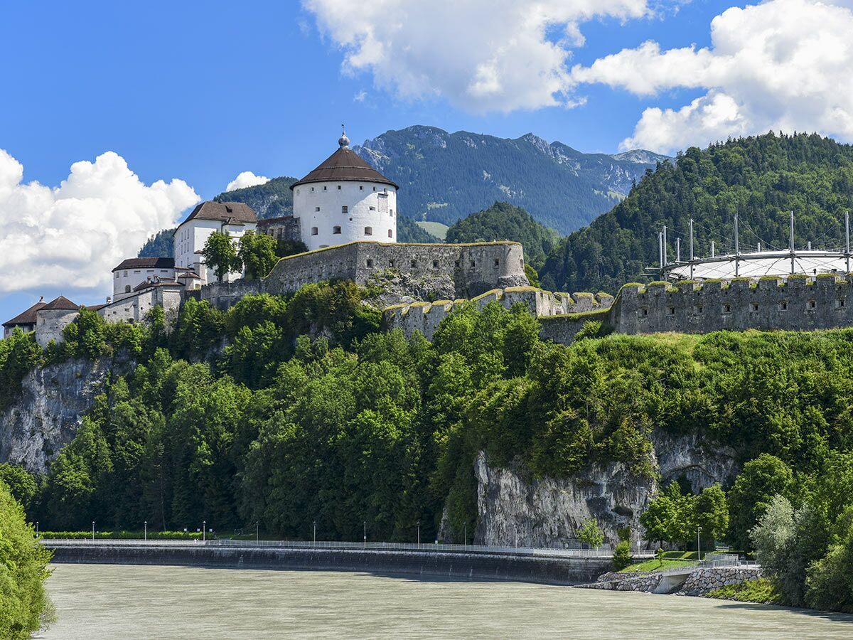 Historische Festung Kufstein auf Felsen über dem Inn in Tirol, umgeben von grünen Bäumen und Bergen unter blauem Himmel.