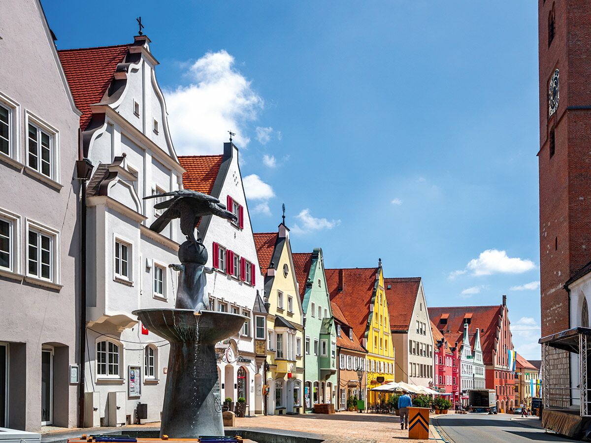Blick auf einen historischen Marktplatz mit einem detaillierten Brunnen mit Vogelstatue, einer Reihe bunter Giebelhäuser und einem Kirchturm unter blauem Himmel.