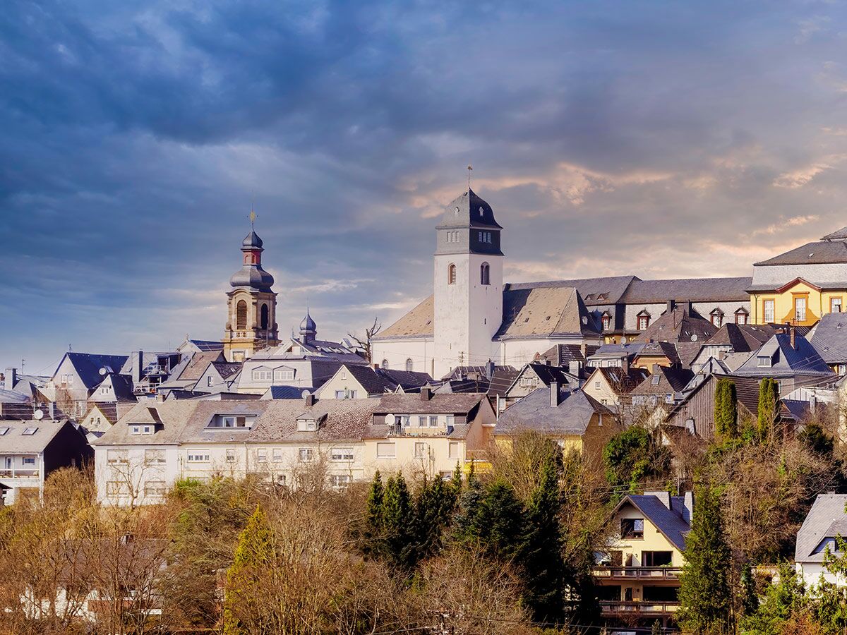Panoramablick auf eine europäische Kleinstadt mit historischen Kirchen und Wohngebäuden auf einem Hügel unter dramatischem Himmel.