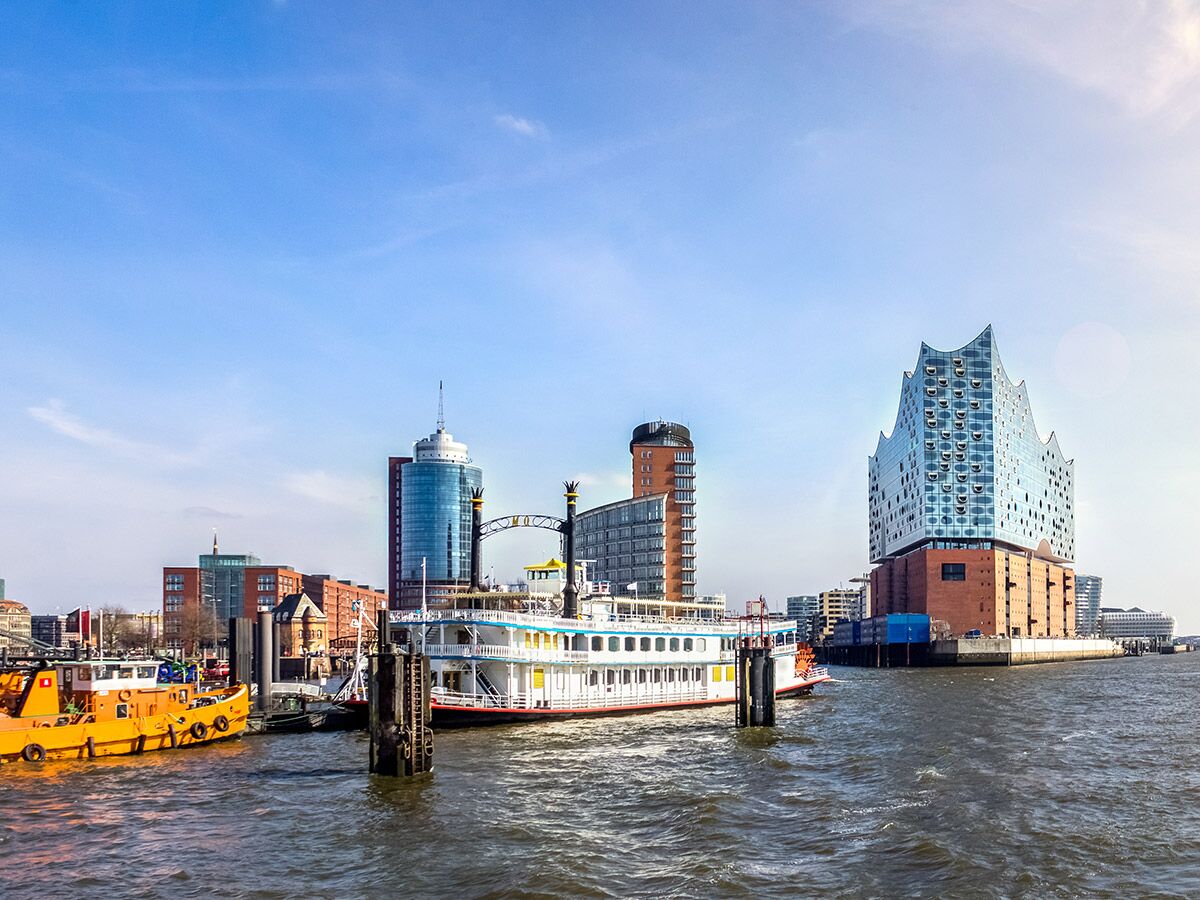 Moderne Elbphilharmonie und andere Gebäude am Elbufer in Hamburg mit Booten auf dem Fluss unter blauem Himmel.