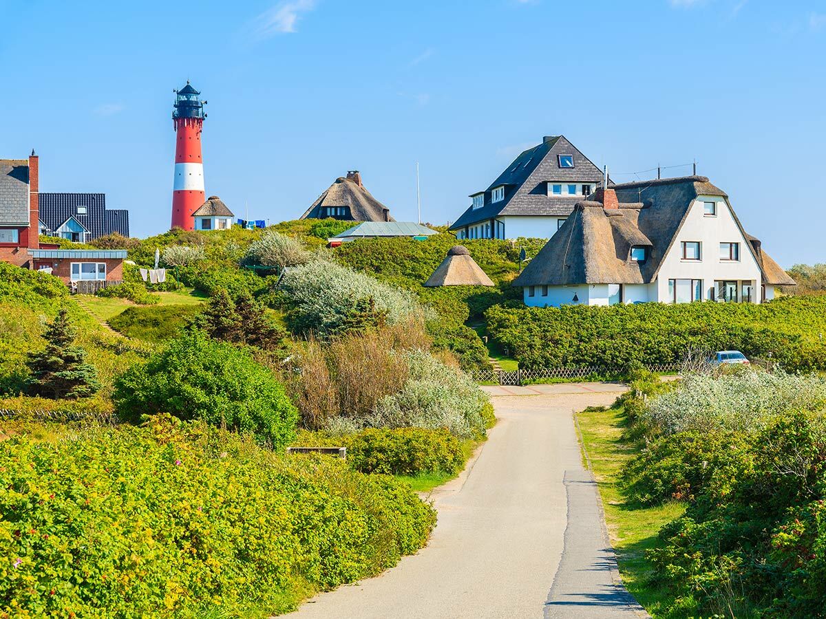 Roter Leuchtturm und traditionelle Reetdachhäuser auf grünen Hügeln einer Nordseeinsel unter blauem Himmel. Idyllische Küstenlandschaft.
