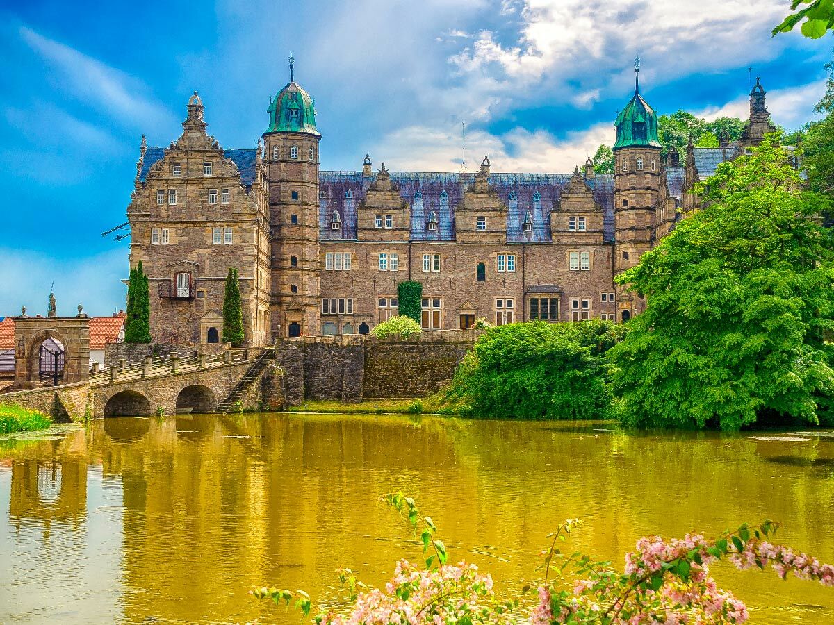 Historisches Wasserschloss mit grünen Dächern und Türmen, umgeben von einem gelb-braunen See und grünen Bäumen unter blauem Himmel.