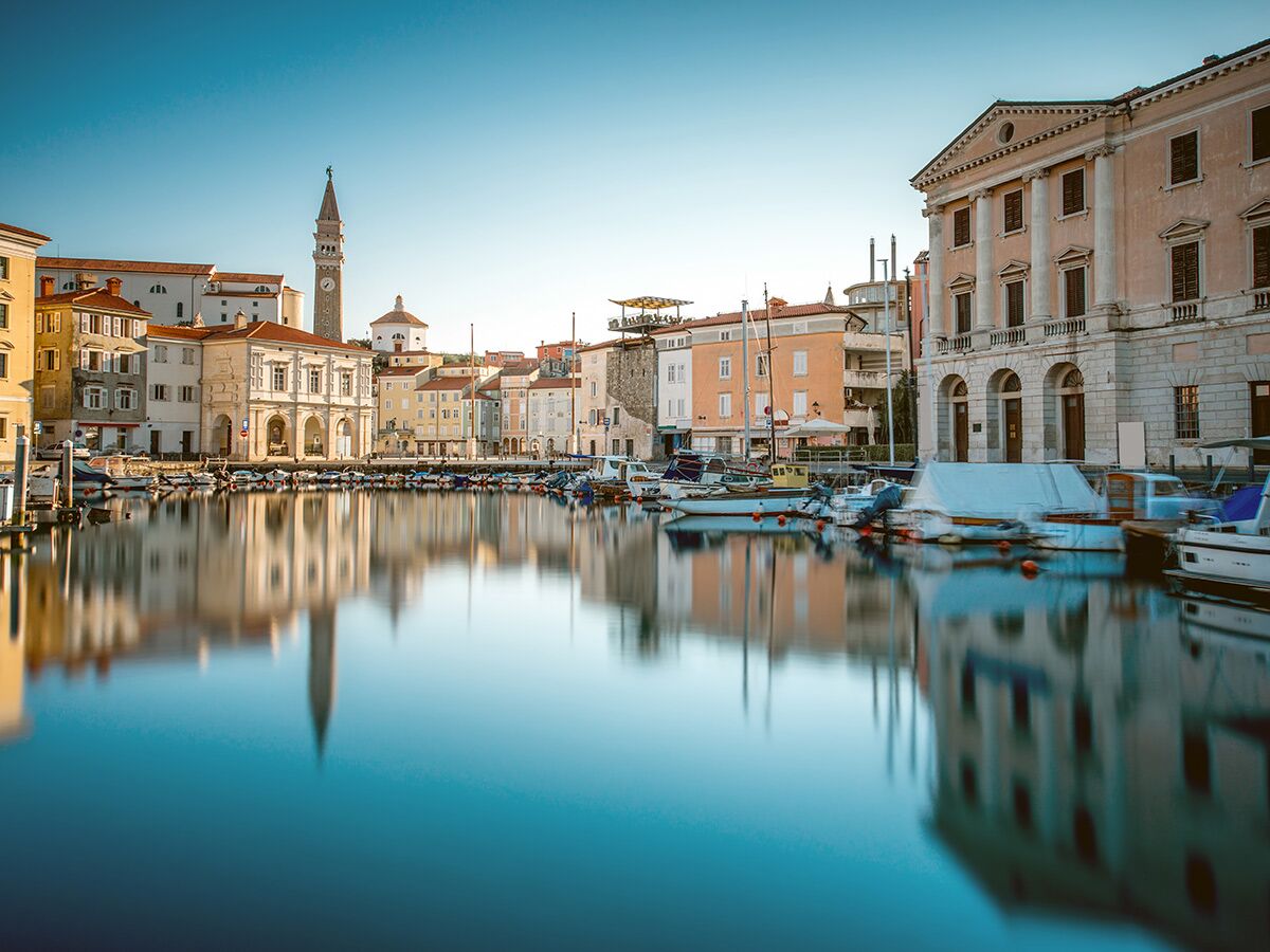 Malerischer Hafen von Piran, Slowenien, mit bunten Häusern, Glockenturm und vielen Booten, die sich im ruhigen Wasser spiegeln.