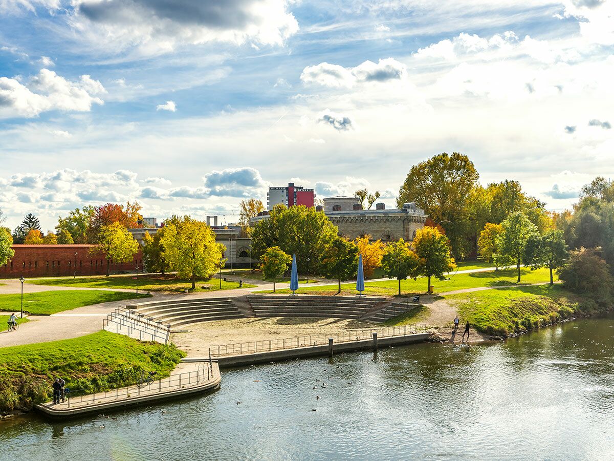 Sonniger Festungspark im Herbst mit farbigen Bäumen, Flussufer, Amphietheater, historischen Gebäuden und Menschen.