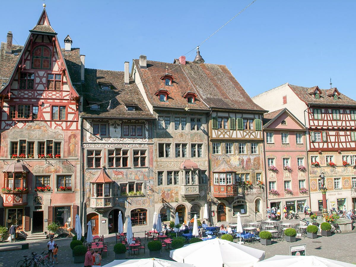 Blick auf einen belebten Marktplatz mit bunten, bemalten Fachwerkhäusern, Außengastronomie, Menschen und Fahrrädern an einem sonnigen Tag.